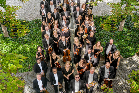 Orchesterbild der Musikerinnen und Musiker der Münchner Symphoniker. Das Bild ist aus der Vogelperspektive aufgenommen, die Orchestermitglieder schauen nach oben in die Kamera. Sie werden von grünen Bäumen umrahmt.