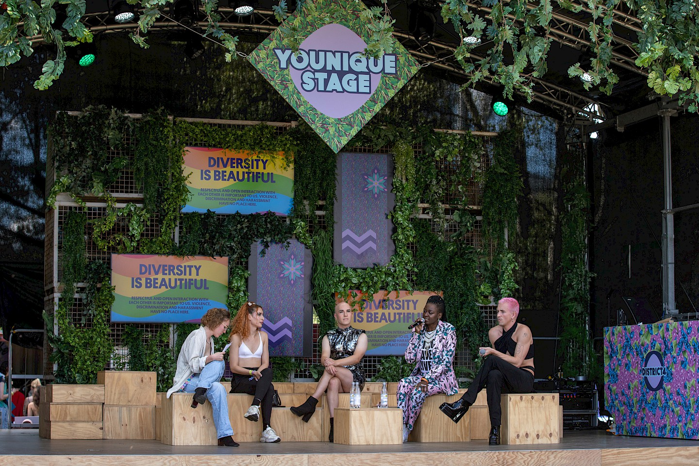 Four people are sitting on a stage and talking in a panel, behind them is a wall covered with green plants on which there is a screen with the text "Diversity is beautiful".