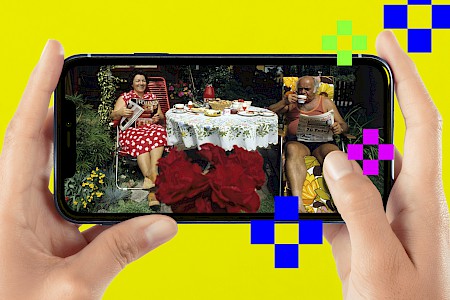 Eine Person hält ein Handy in der Hand. Darauf ist ein Foto zu sehen mit dem Titel „Schrebergartenidylle“. Ein Ehepaar sitzt in Liegestühlen an einem gedeckten Tisch in seinem Kleingarten.