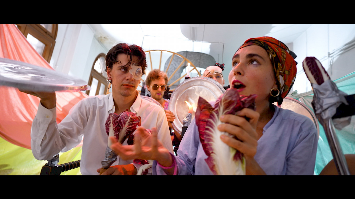 A colorful collective is shown in an artistic performance in a bright room. In the foreground are a young man and a young woman who are disguised and look like a pirate and a pirate woman with their golden earrings and headscarf. The man is holding a tin sign and a radicchio salad, just as the woman is holding a radicchio salad. She looks up, waves her right hand and calls out something.