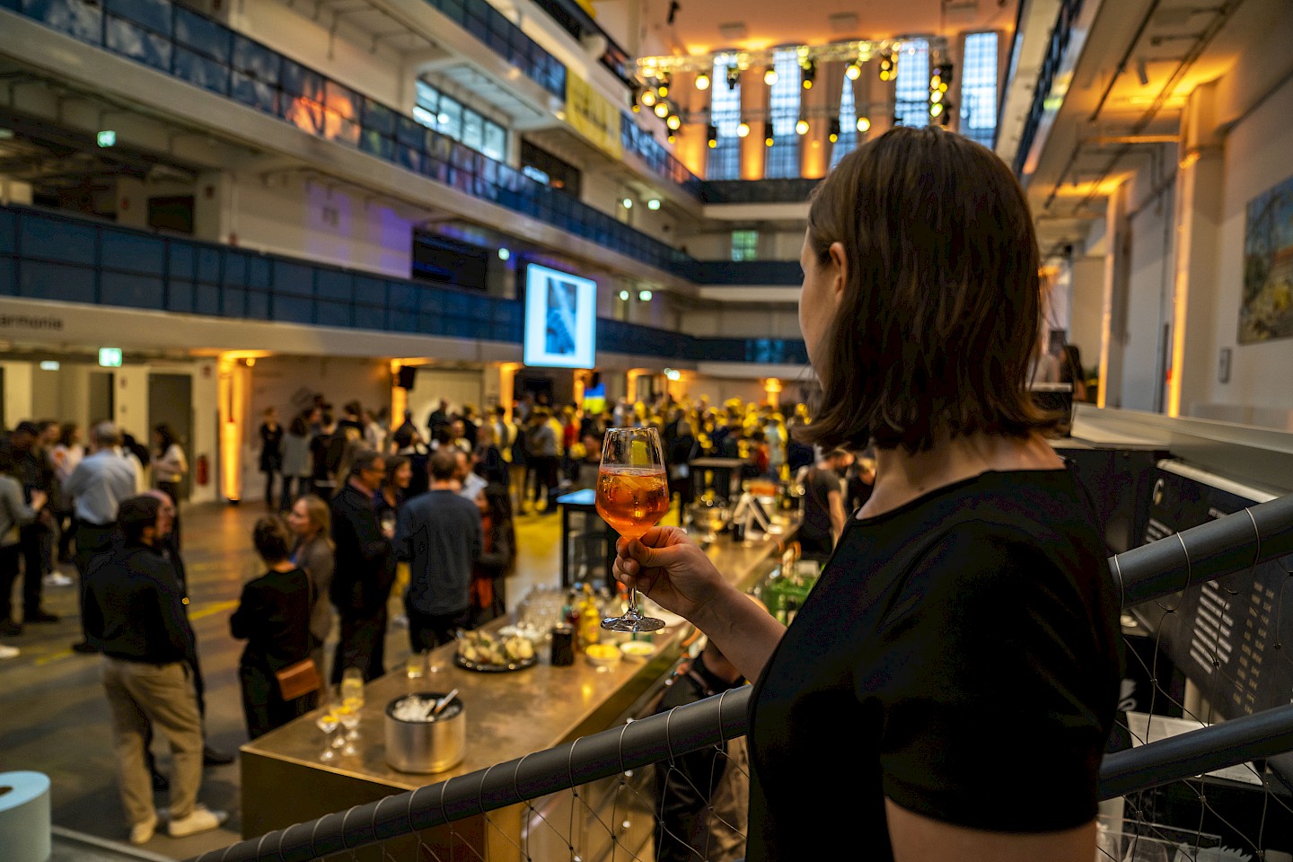 In the foreground, a woman with drink in hand, looking at a reception in the hall in the background.