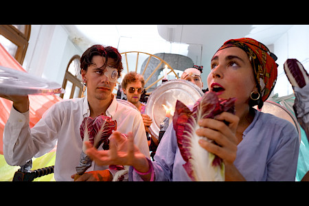 A colorful collective is shown in an artistic performance in a bright room. In the foreground are a young man and a young woman who are disguised and look like a pirate and a pirate woman with their golden earrings and headscarf. The man is holding a tin sign and a radicchio salad, just as the woman is holding a radicchio salad. She looks up, waves her right hand and calls out something.